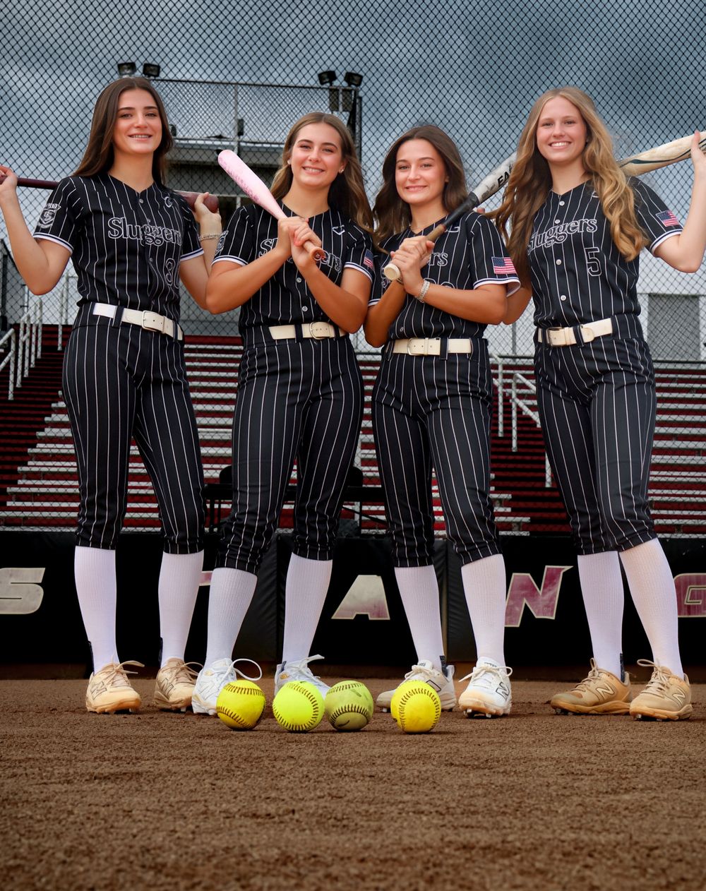 Four female softball players in black pinstripe uniforms pose with bats and softballs on a dirt field.
