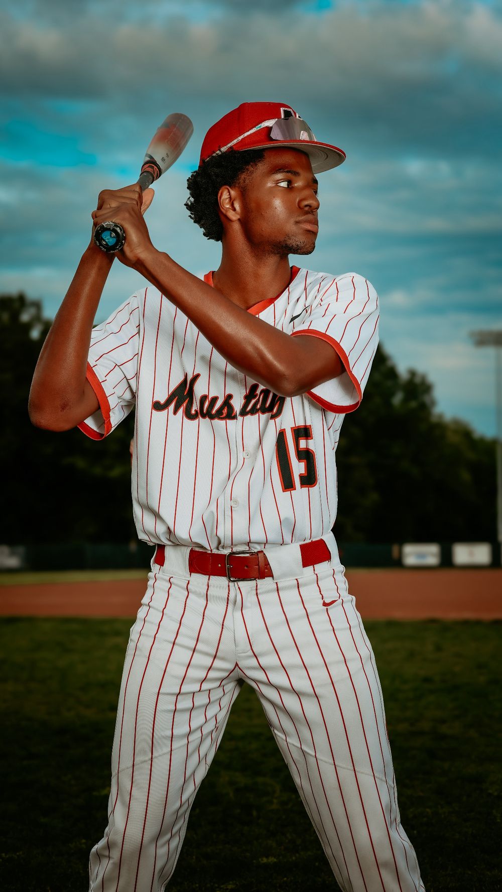 Black teen baseball player in Mustangs number 15 uniform holds bat in batting stance on field.