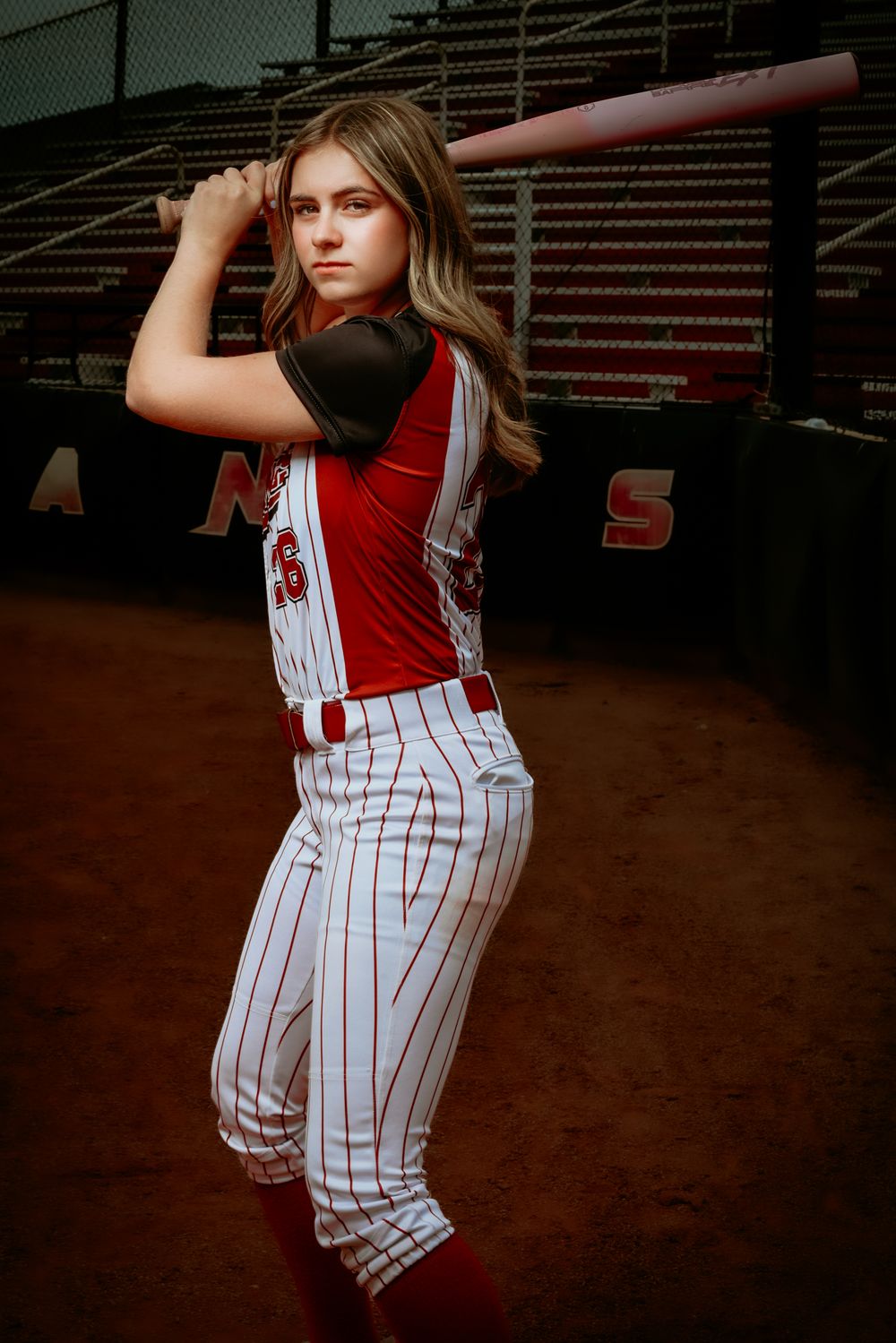 Young female softball player in red and white pinstripe uniform poses with pink bat at stadium.
