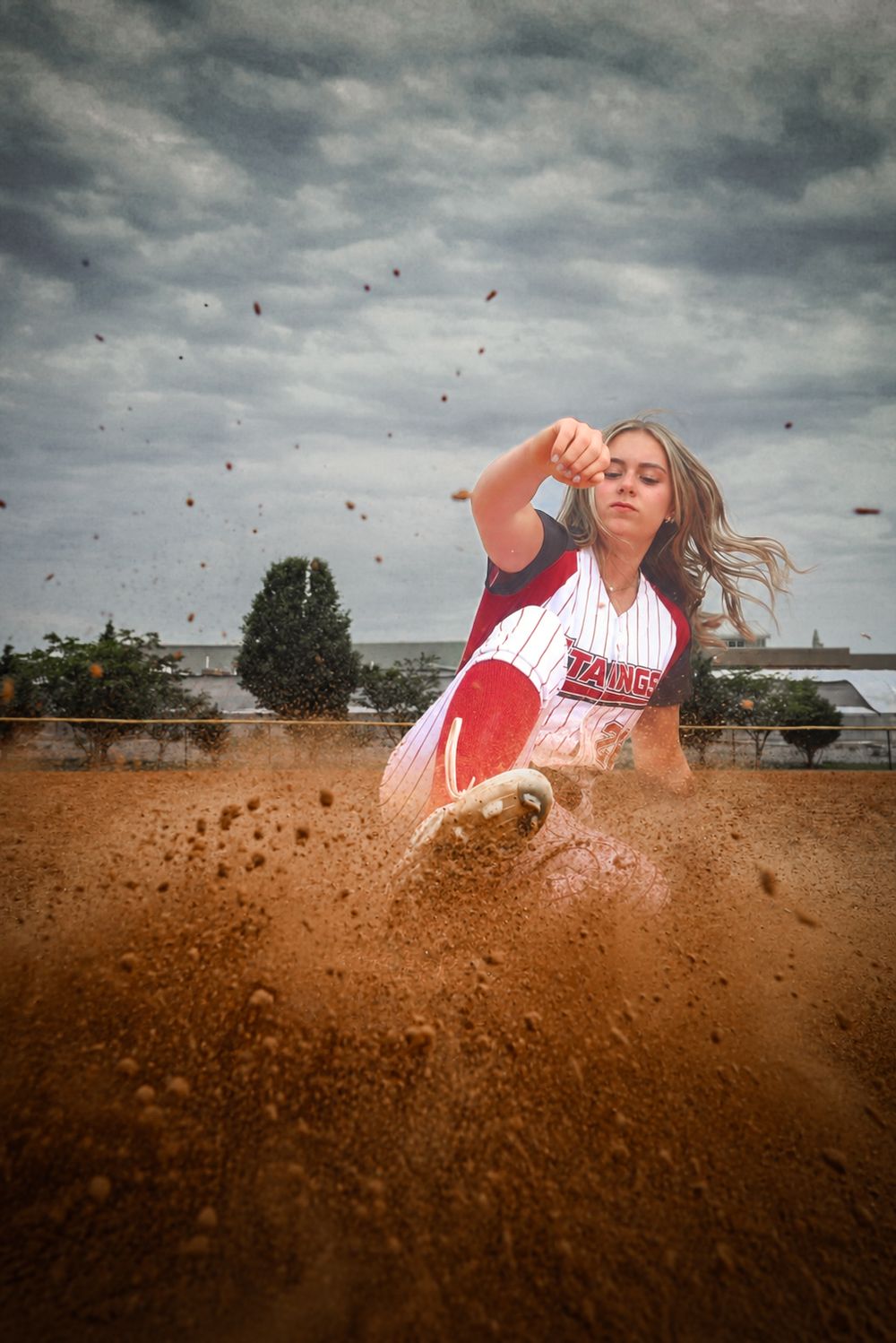 Female softball player sliding into base on dirt field, wearing red and white uniform, with dirt flying dramatically under cloudy sky.