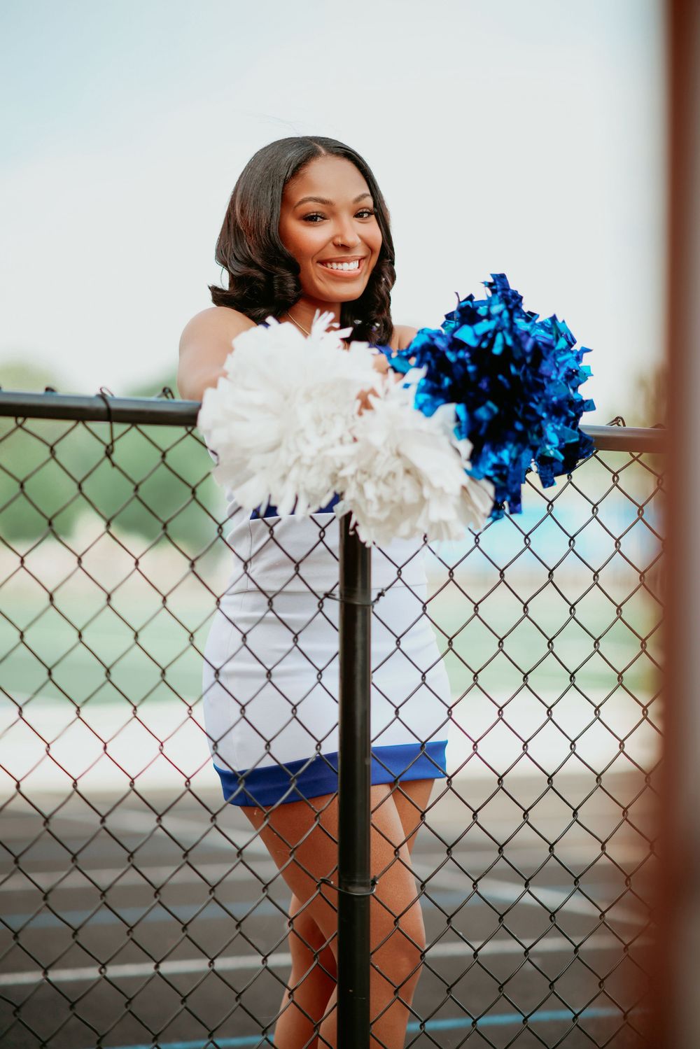 Smiling Black cheerleader in blue and white uniform holds pom-poms behind a chain-link fence.