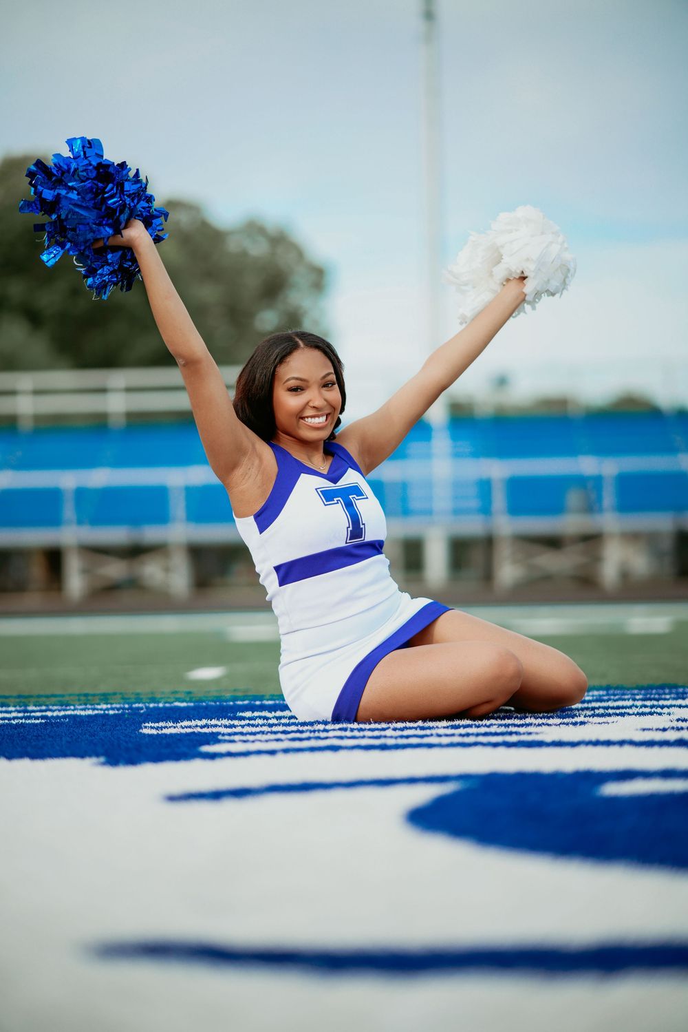 Black cheerleader in blue and white uniform kneels on football field with pom-poms raised overhead.