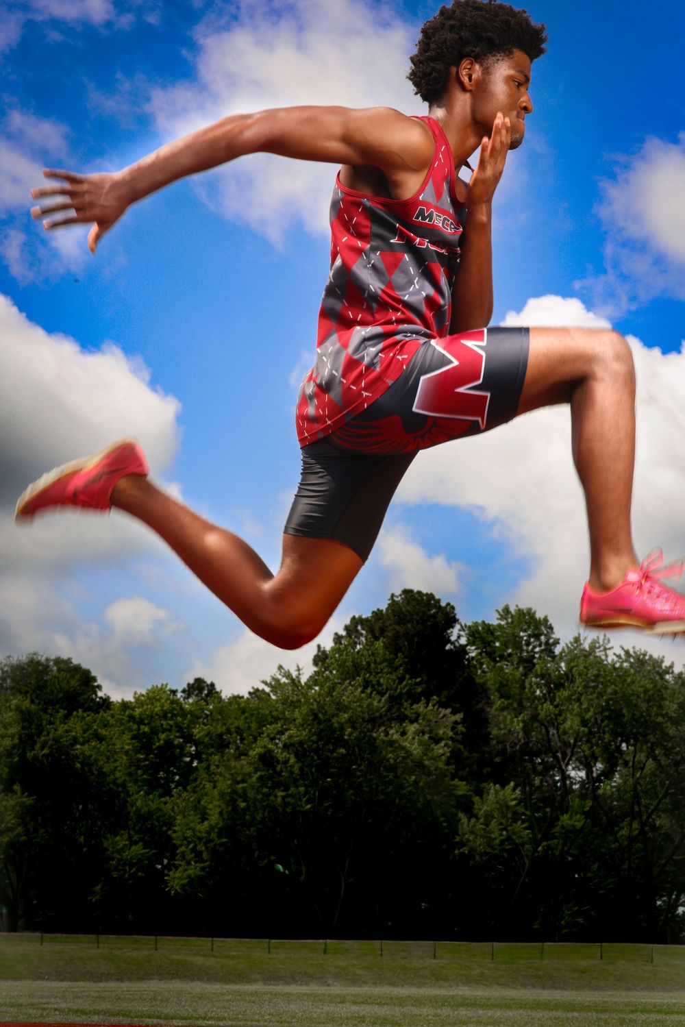 Male track athlete in red Mustangs uniform leaping dynamically against a bright blue cloudy sky.
