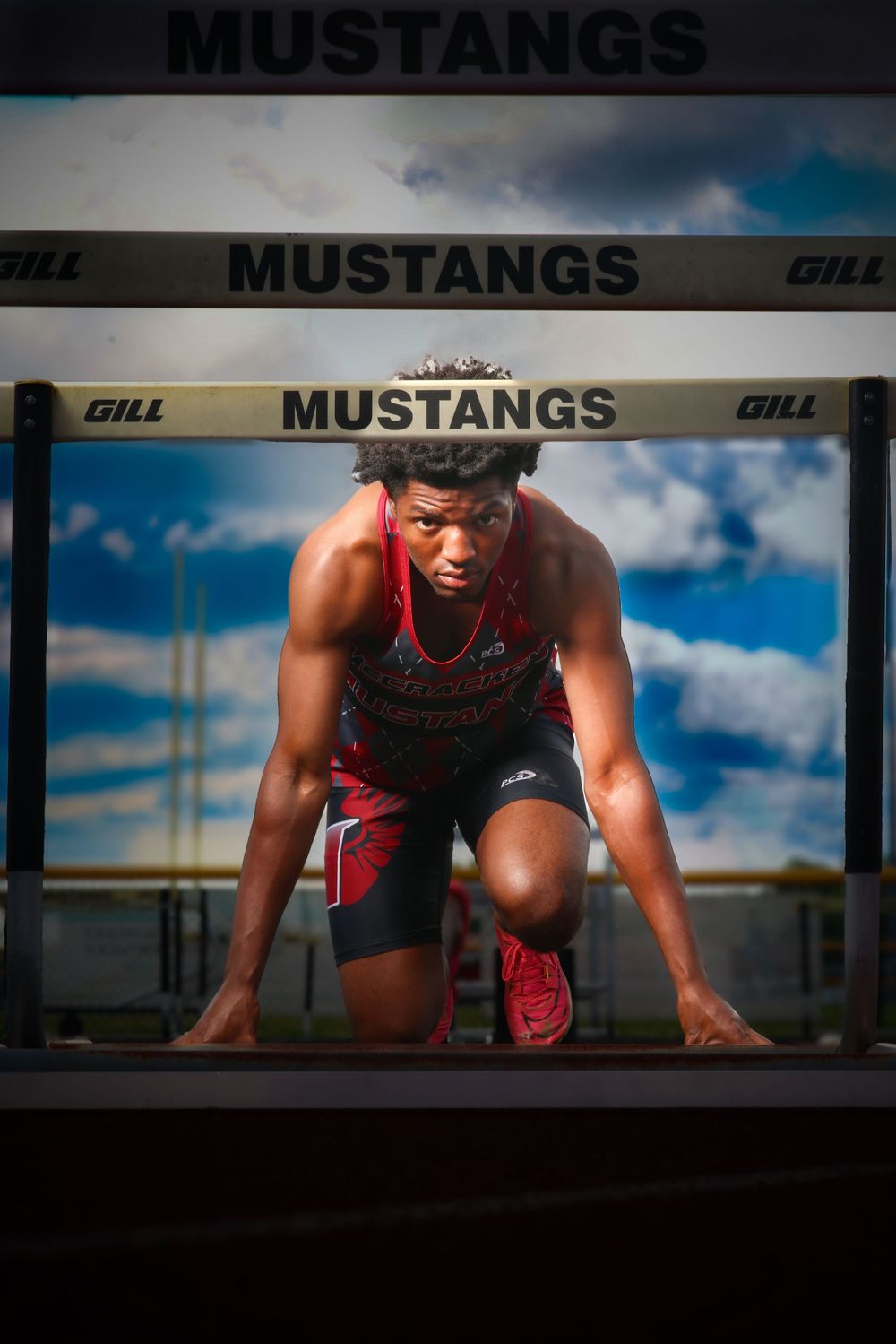 Male track athlete in starting position beneath Mustangs hurdles with dramatic cloudy sky background.