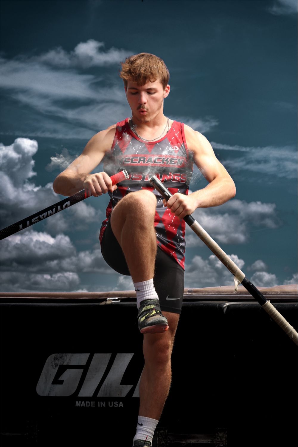 Young male pole vaulter in red athletic uniform grips a Gill pole, preparing for his vault under a dramatic cloudy sky.