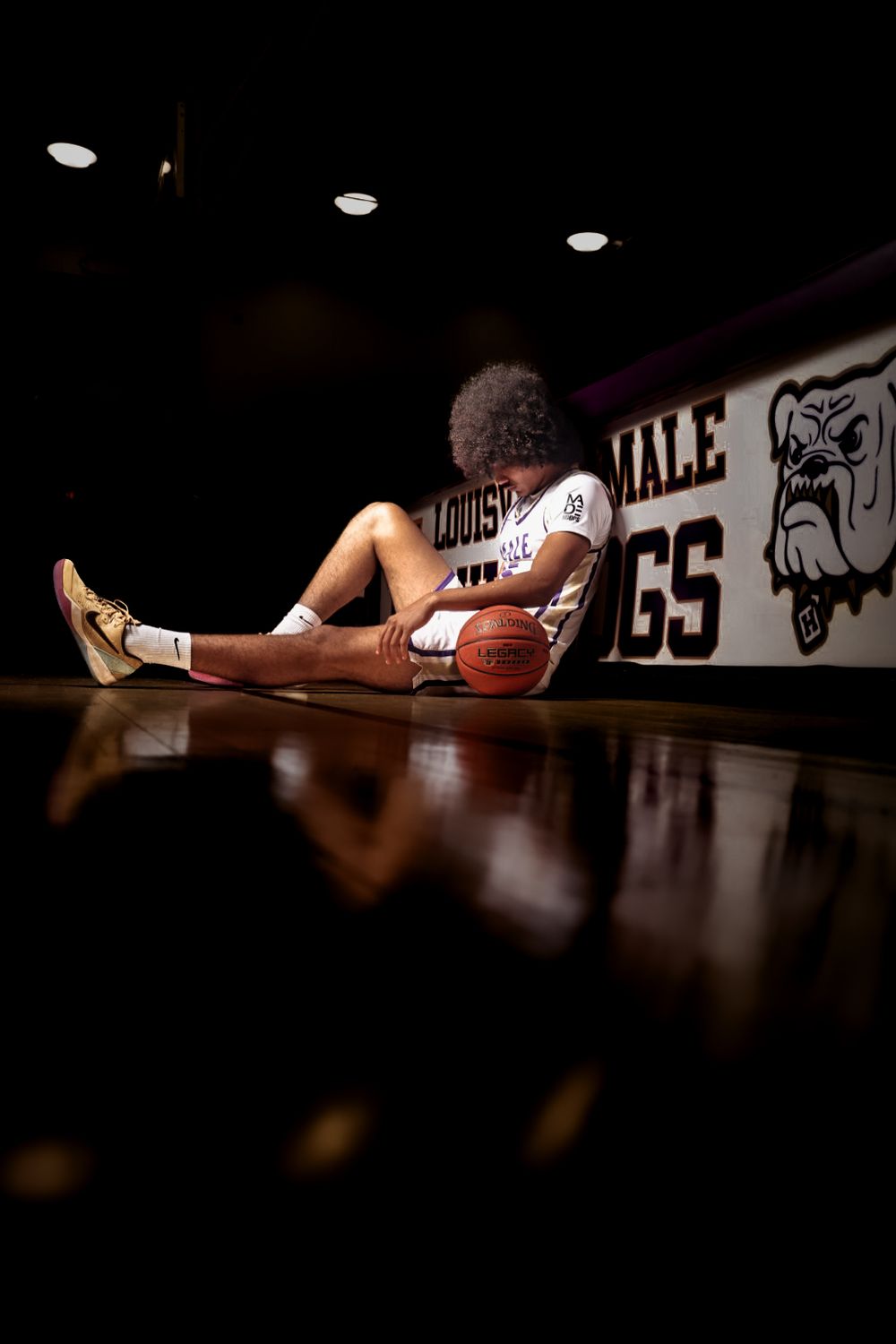 Basketball player sitting on court floor holding ball, leaning against Louisville Male Bulldogs gym wall in white jersey.