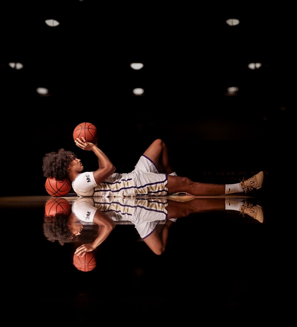 Basketball player in white uniform lying on reflective court floor holding ball, dramatic dark arena lighting.