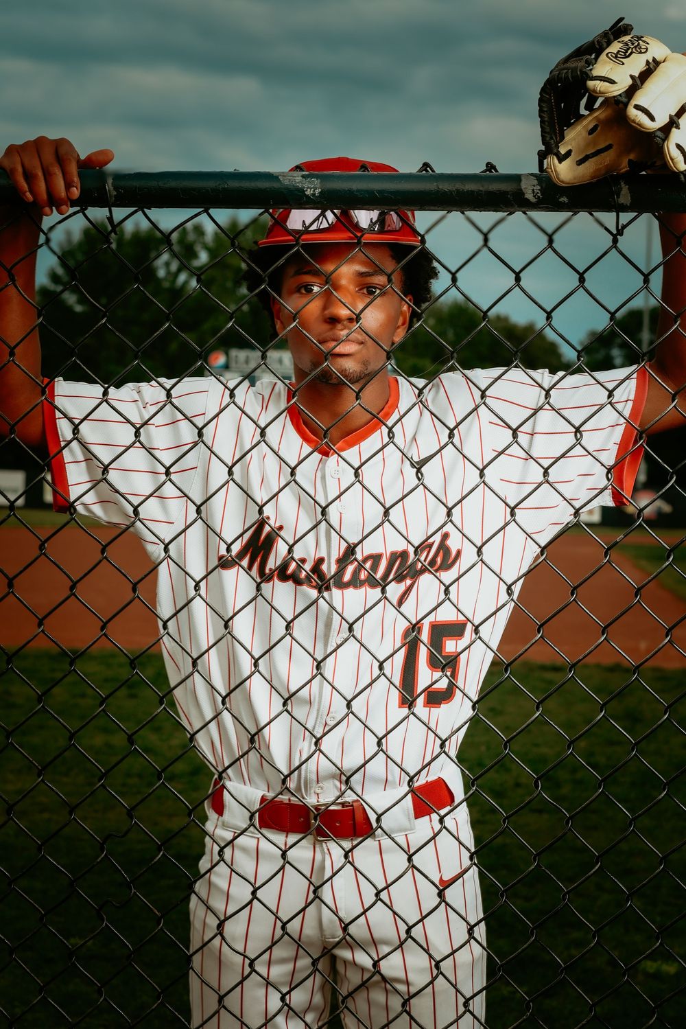 Black teen baseball player in Mustangs number 15 uniform leans against chain-link fence on field.