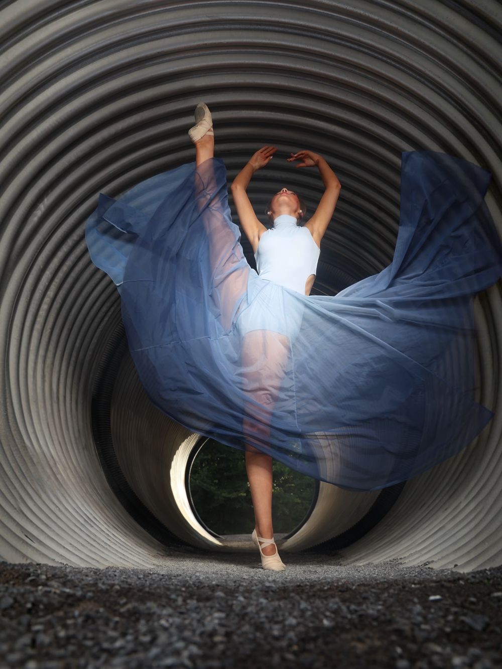 Ballerina in white tutu poses gracefully inside a large metal tunnel, blue fabric flowing dramatically around her.