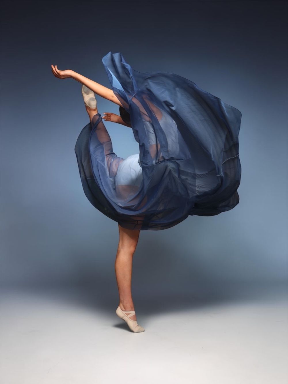 Ballerina in flowing blue sheer dress performing elegant arabesque on pointe in a studio.