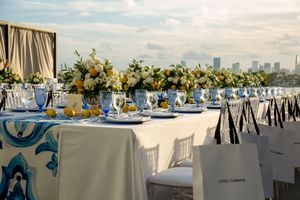 Elegant waterfront wedding reception setup with white tables, blue accents, and city skyline views at sunset.