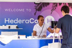 A woman staffing a registration desk at HederaCon event with a colorful welcome banner backdrop.