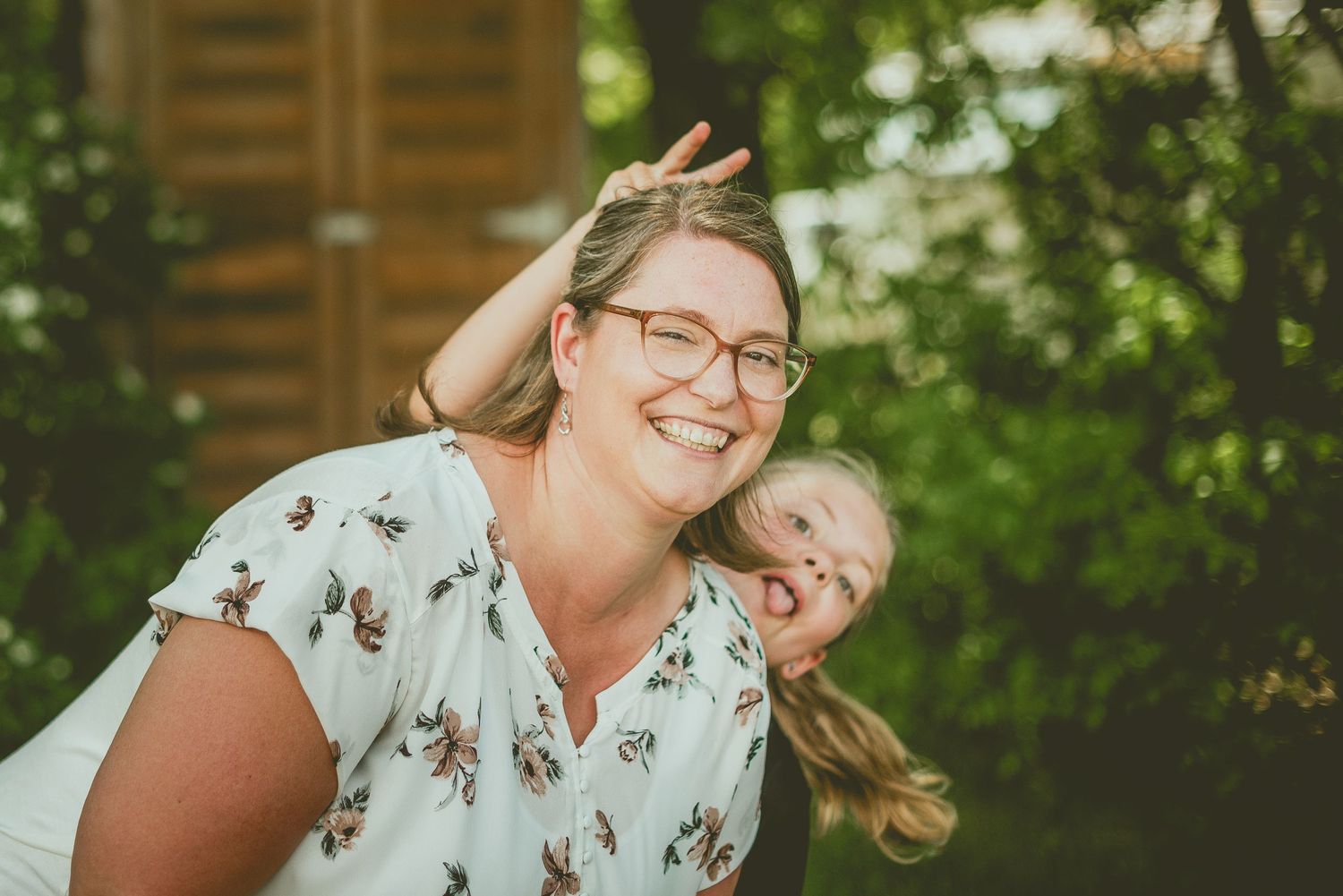 A laughing woman with glasses and a floral top plays outdoors with a child in a lush green garden setting.