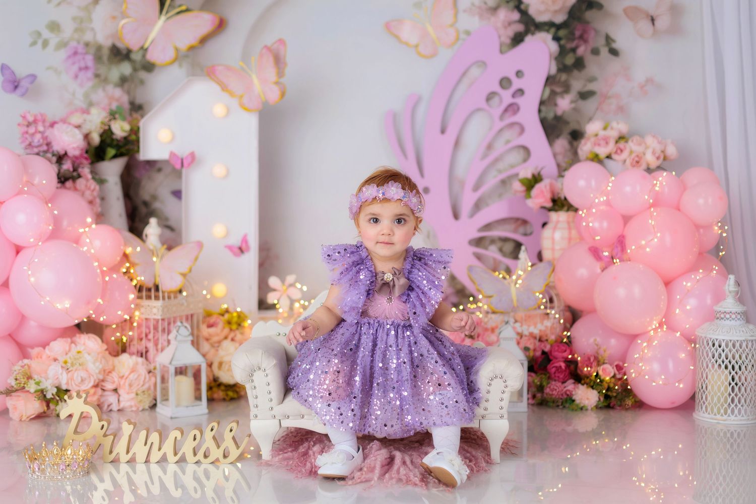 Baby girl in purple dress sits on white chair with butterfly wings, pink balloons, and Princess sign at first birthday party.