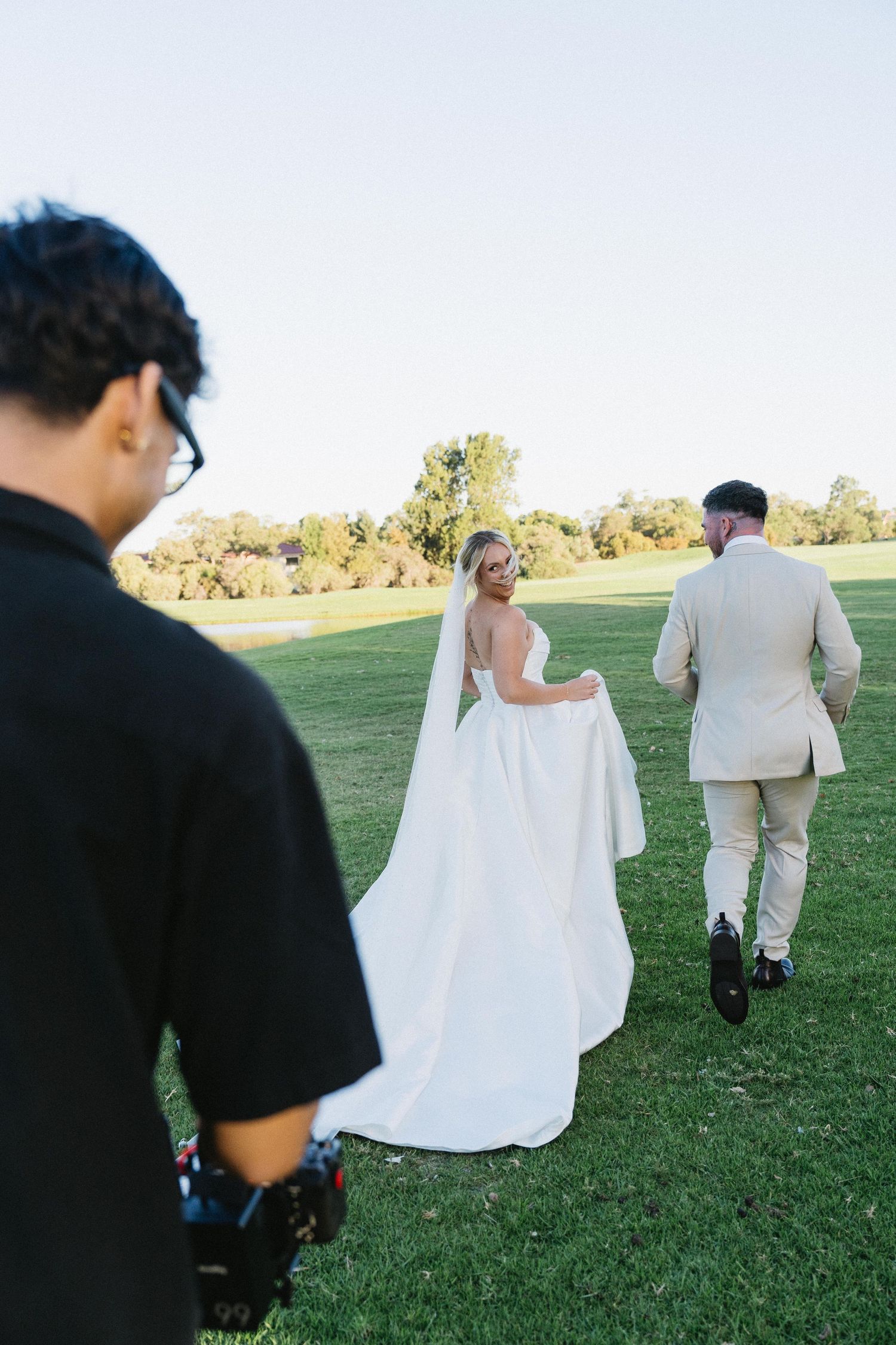 Videographer filming bride in white gown and groom in beige suit walking on lush green lawn at outdoor wedding.