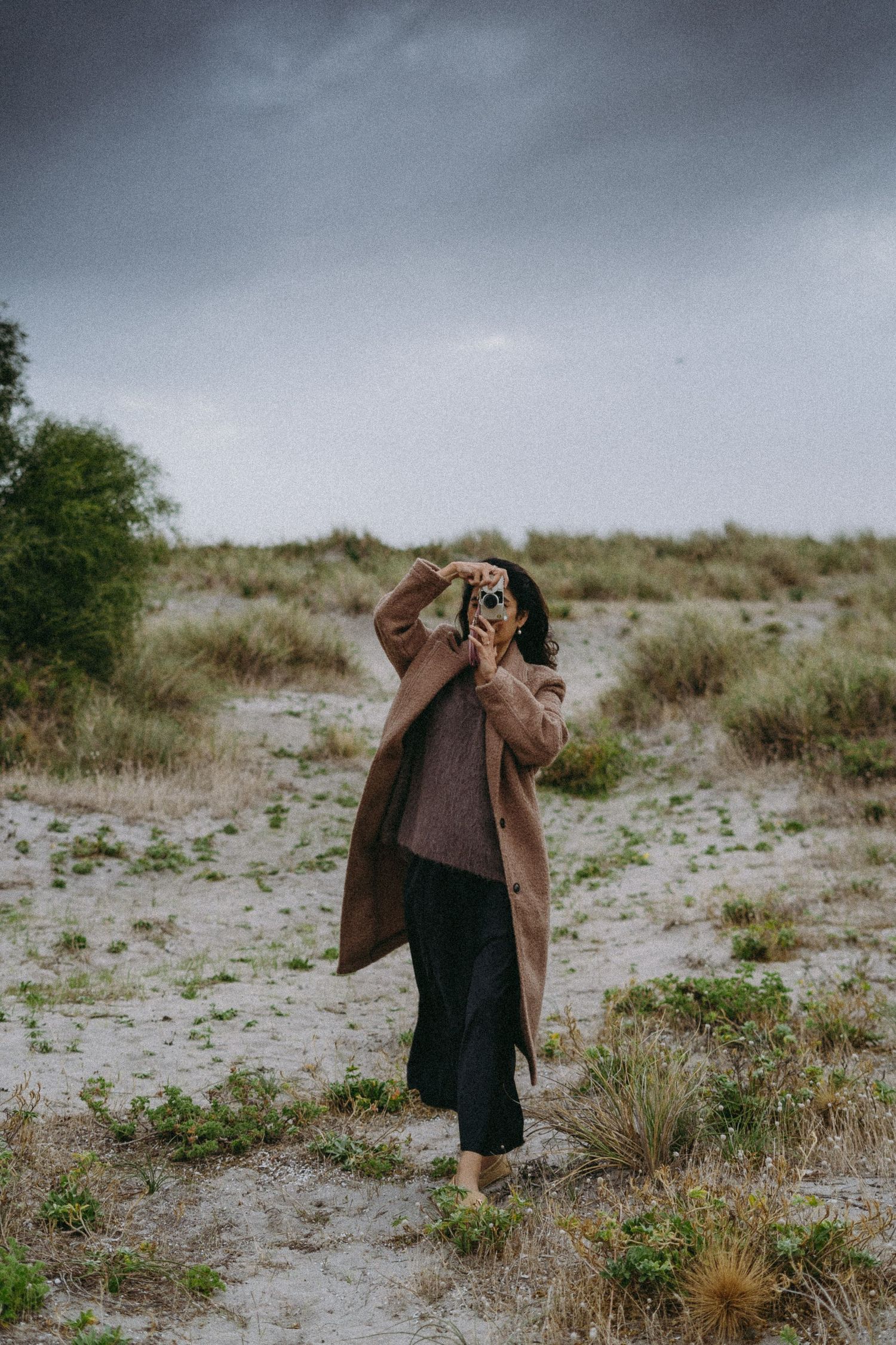 Woman in beige coat shielding eyes from wind on sandy dunes, holding camera, overcast sky background.