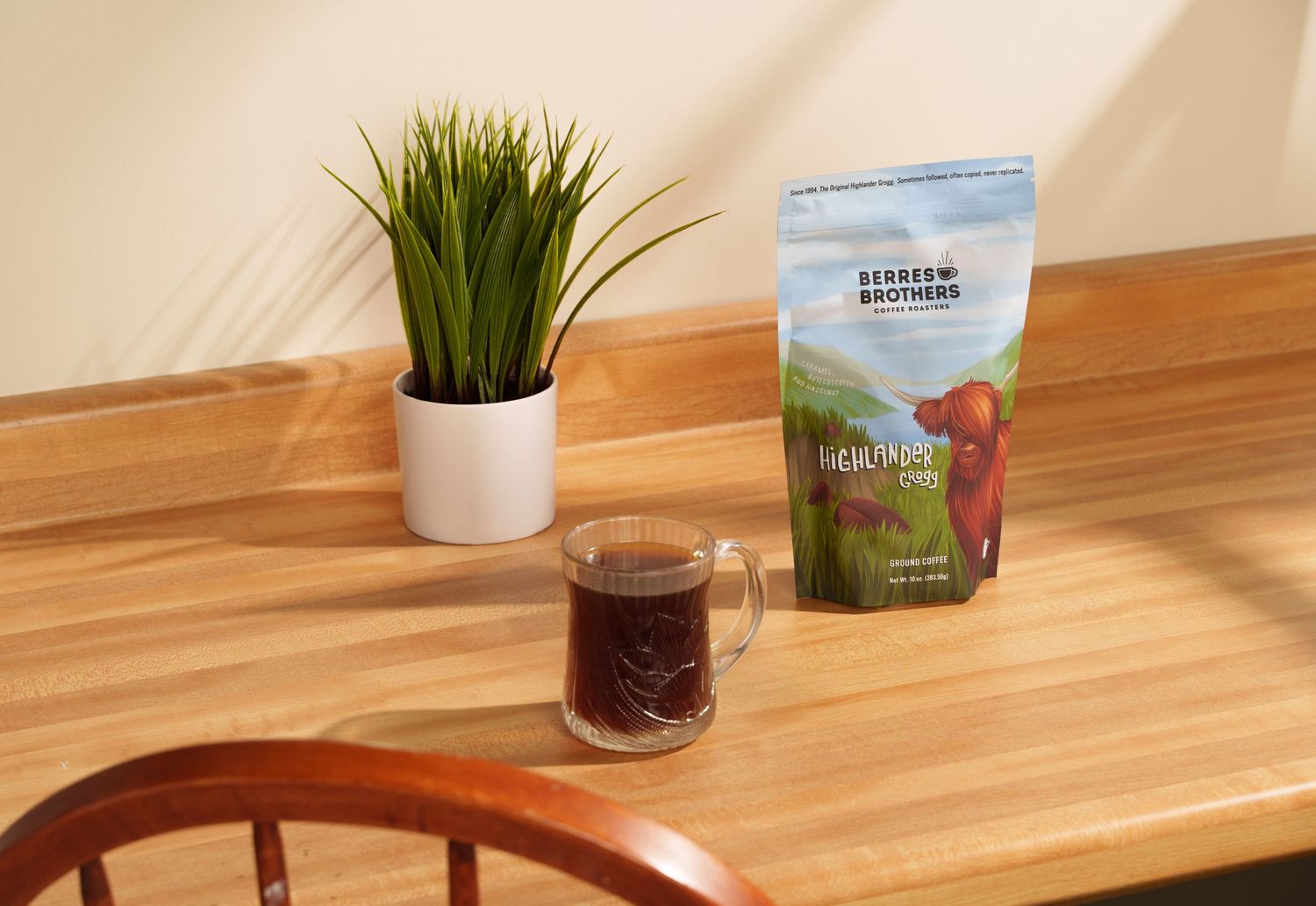 A glass of coffee sits on a wooden table alongside a coffee bag and small potted plant.