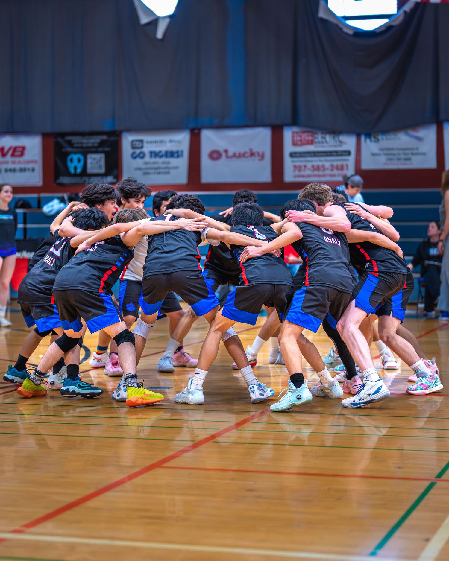 Volleyball team players huddle together in a tight circle during a game in an indoor gymnasium with sponsor banners visible.