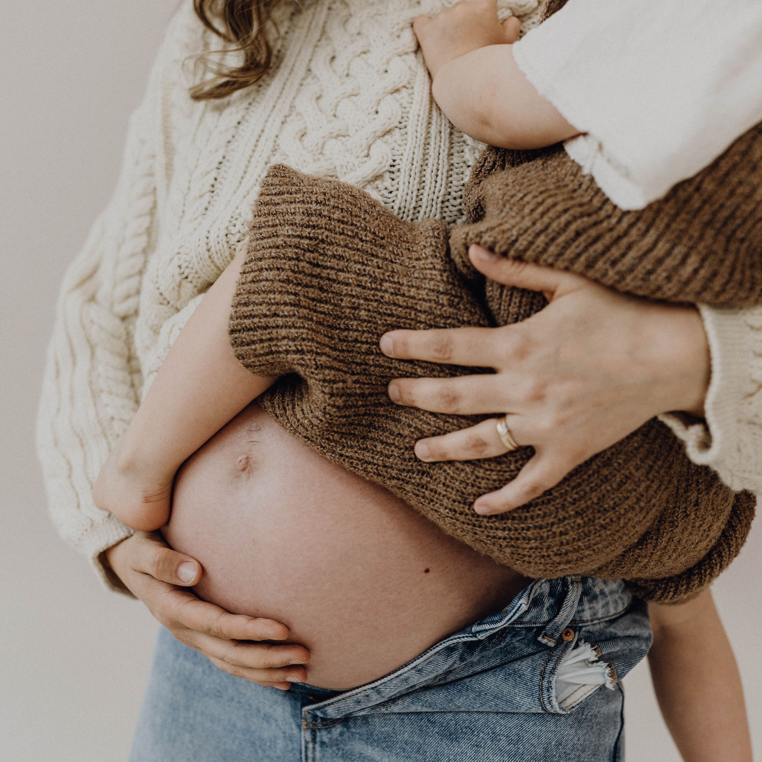 Babybauchshooting im Fotostudio, die Schwangere trägt einen beigefarben Strickpullover, der Bauch ist sichtbar. Auf dem Arm hät sie ein Kind
