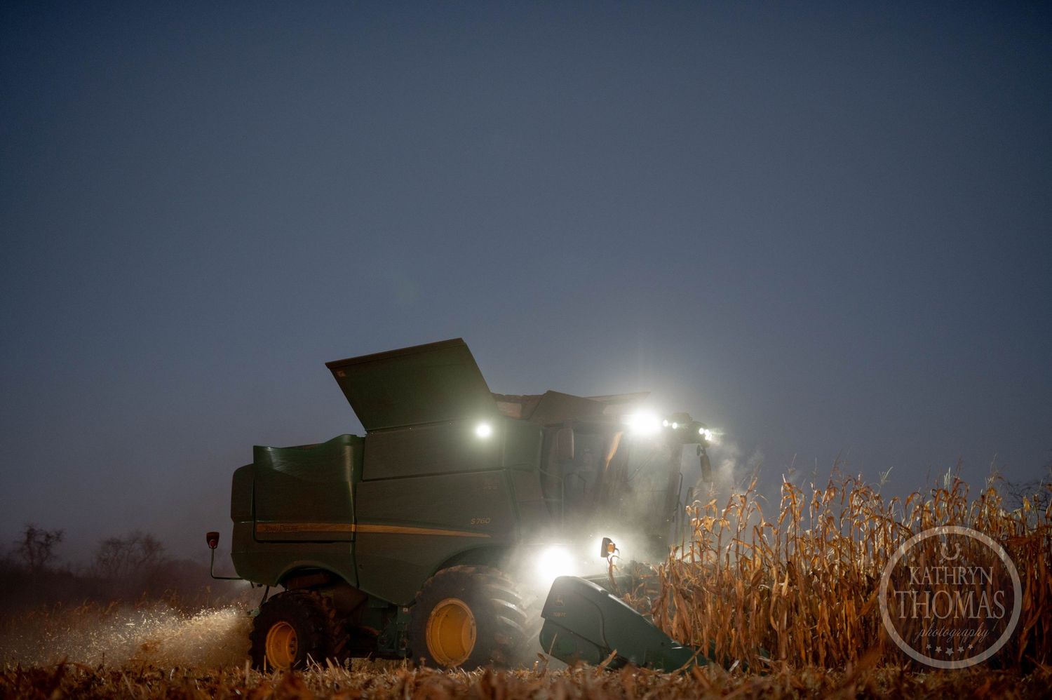 Illuminated combine harvester working in dark cornfields at night.