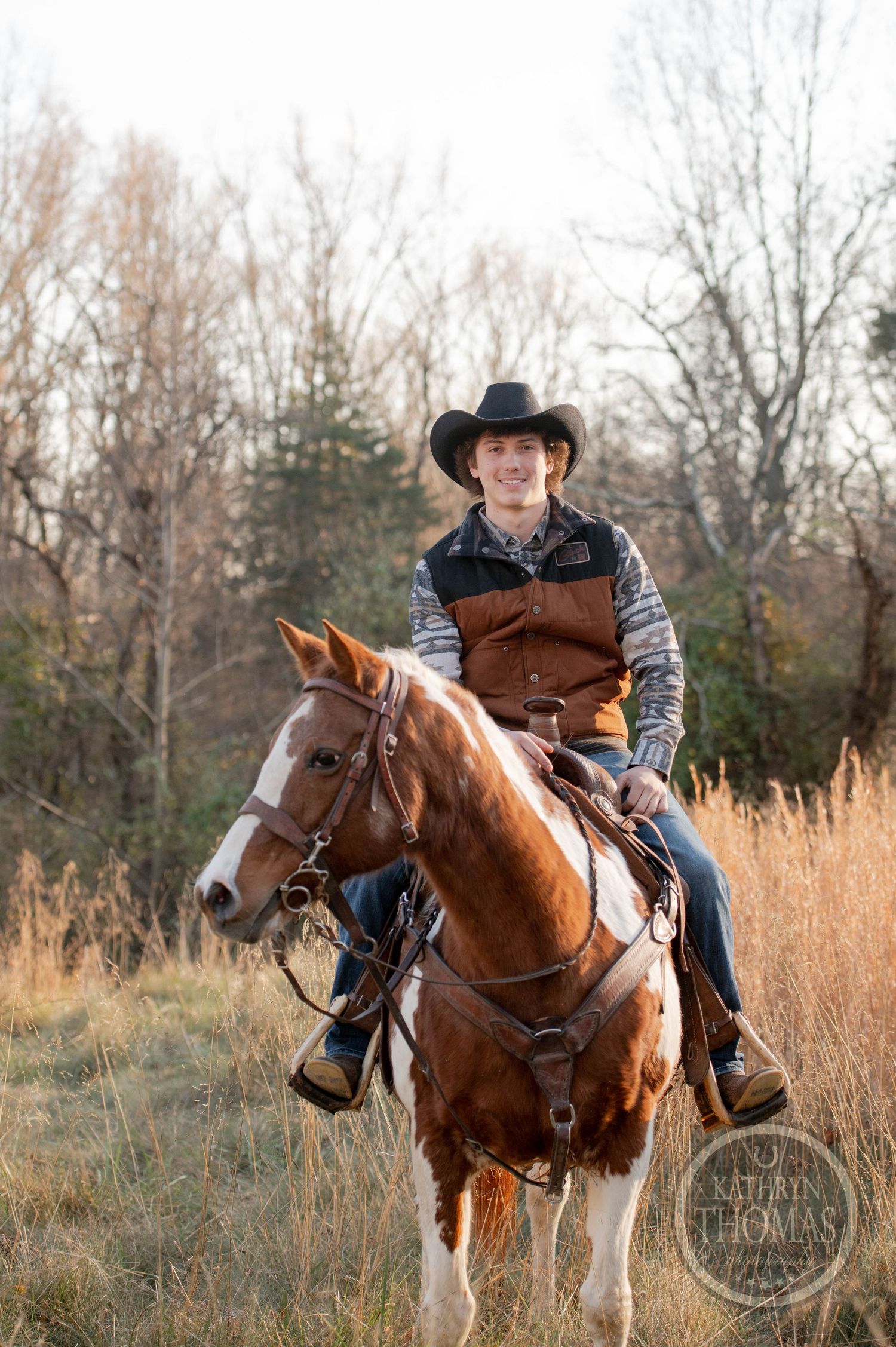 Cowboy on a brown and white horse poses in autumn field with bare trees in background.