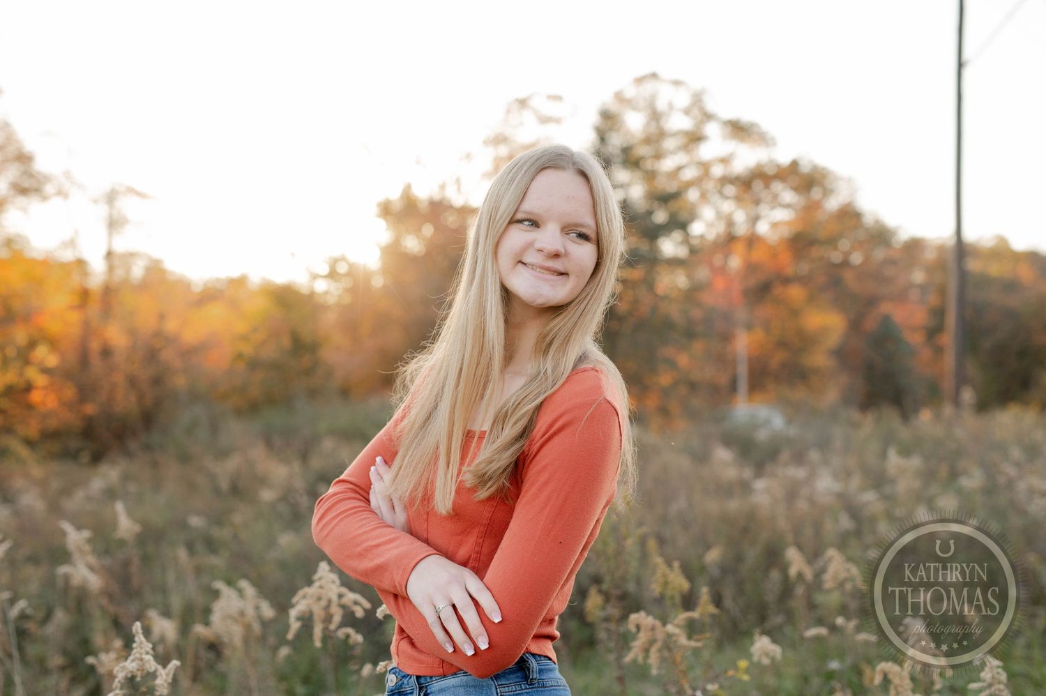 Person in orange long sleeve top poses confidently with arms crossed in fall setting.