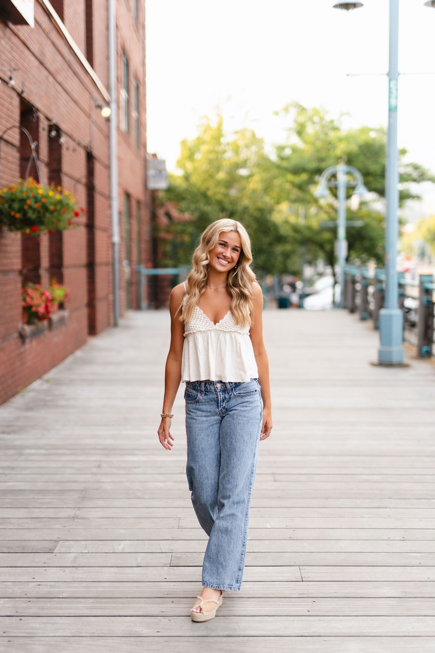A person in a black tank top and white wide-leg pants walks confidently along a wooden boardwalk on a sunny day.