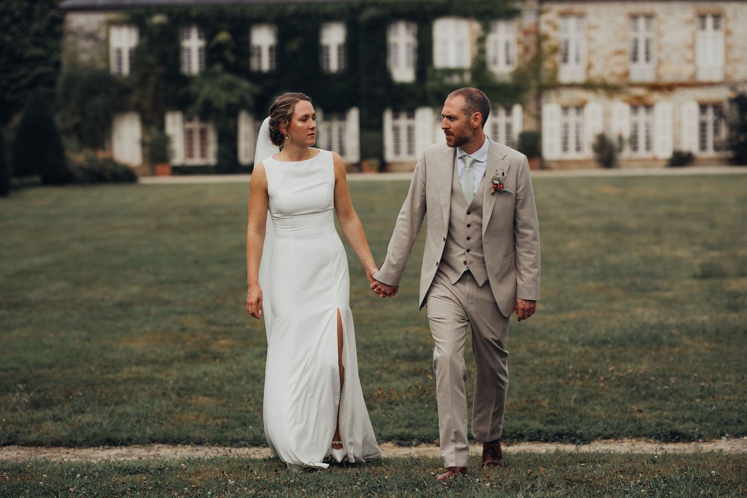 Bride and groom walk hand in hand across green lawn before an elegant ivy-covered chateau on their wedding day.