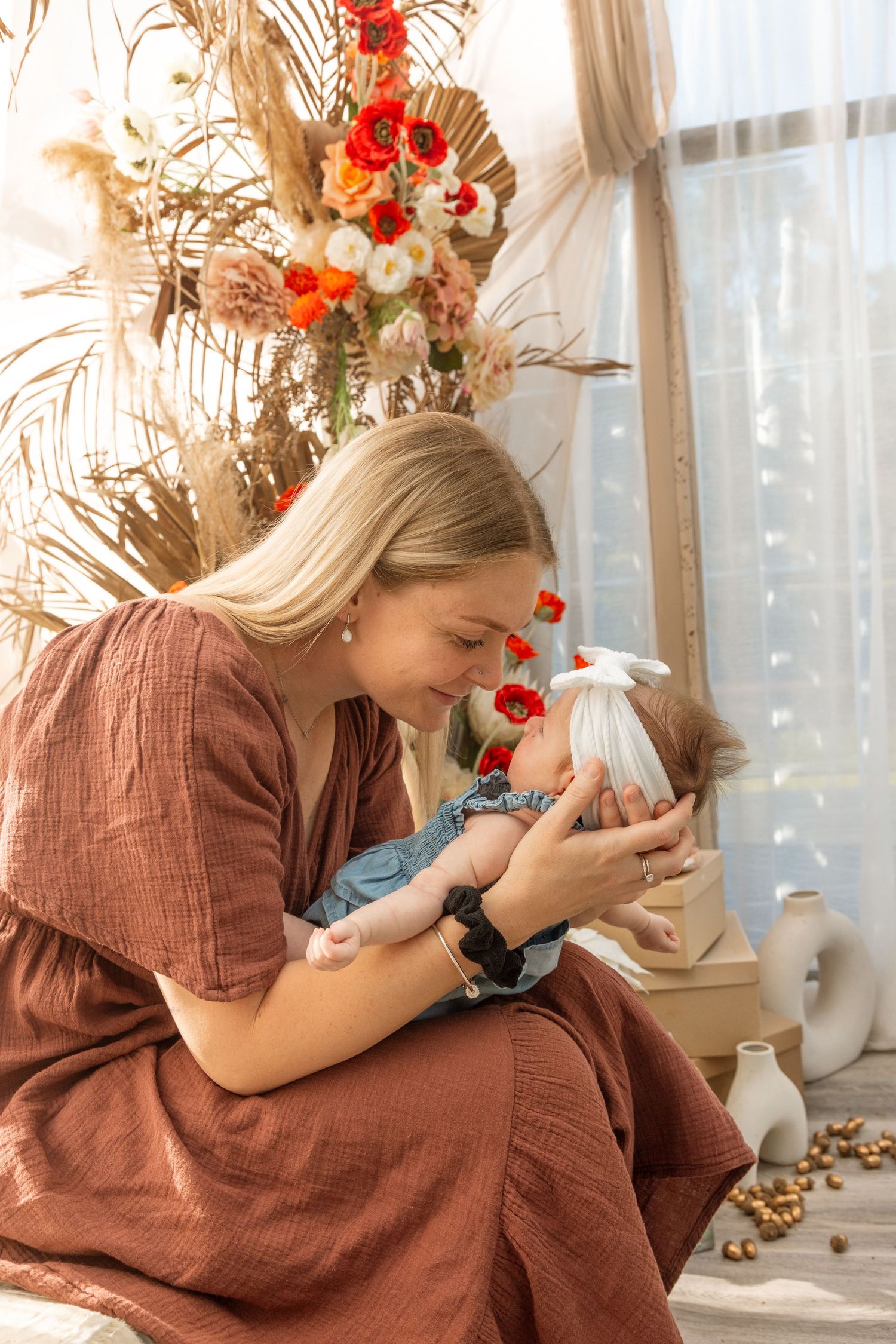 Mother in brown dress tenderly embraces baby in front of floral arrangement and window with dreamy lighting.
