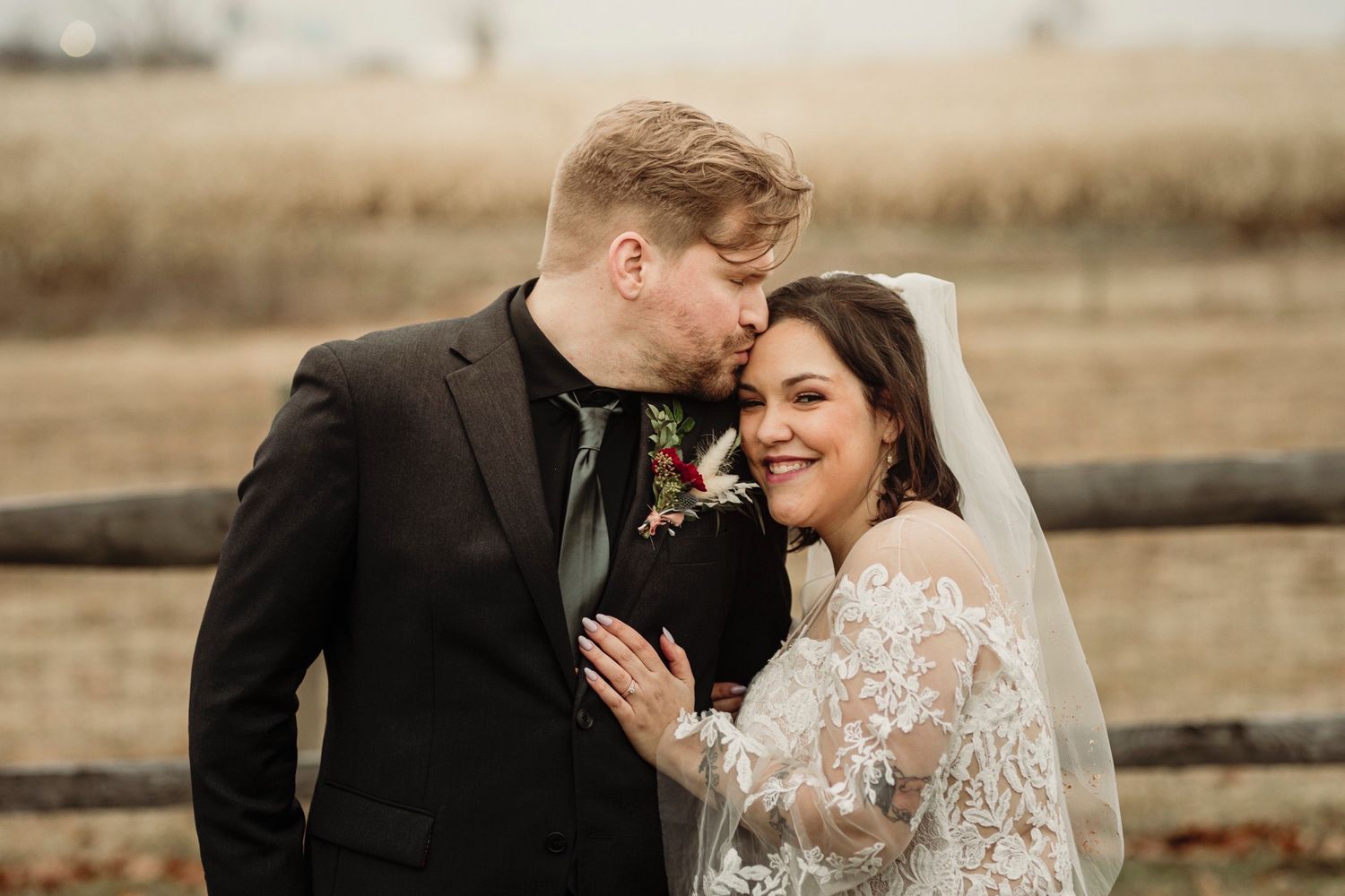 A romantic newlywed couple embraces by a rustic wooden fence in autumn, bride wearing lace and groom in black suit.