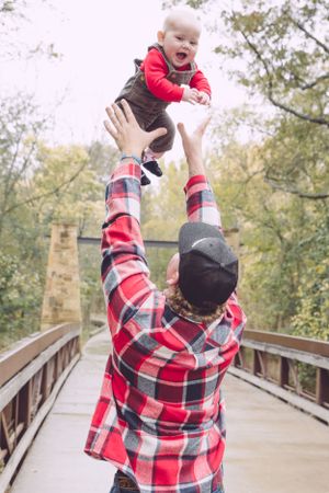 Person in plaid shirt tossing joyful baby in air on wooden bridge.