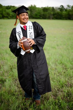 Graduate in cap and gown holds football while posing in grassy field.