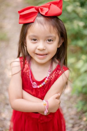 Child in red dress with matching bow accessory poses outdoors.