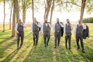 A group of groomsmen in matching gray vests and pants walk together on a sunlit lawn with trees in the background.