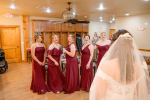 Bridesmaids in burgundy floor-length gowns gather in rustic wooden room during wedding preparations.