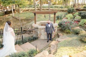 Two people stand in a scenic garden setting with steps and metal railings during an outdoor wedding ceremony.
