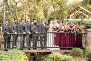 A wedding party in burgundy dresses and gray suits poses together on a wooden bridge in a garden setting.