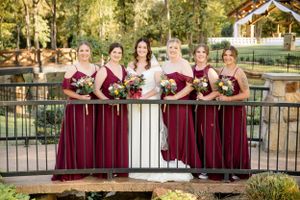 Wedding party group wearing burgundy bridesmaid dresses poses on rustic bridge with gazebo in background during outdoor ceremony.