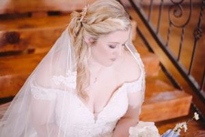 A bride in a white gown and veil looks down while sitting on wooden stairs.