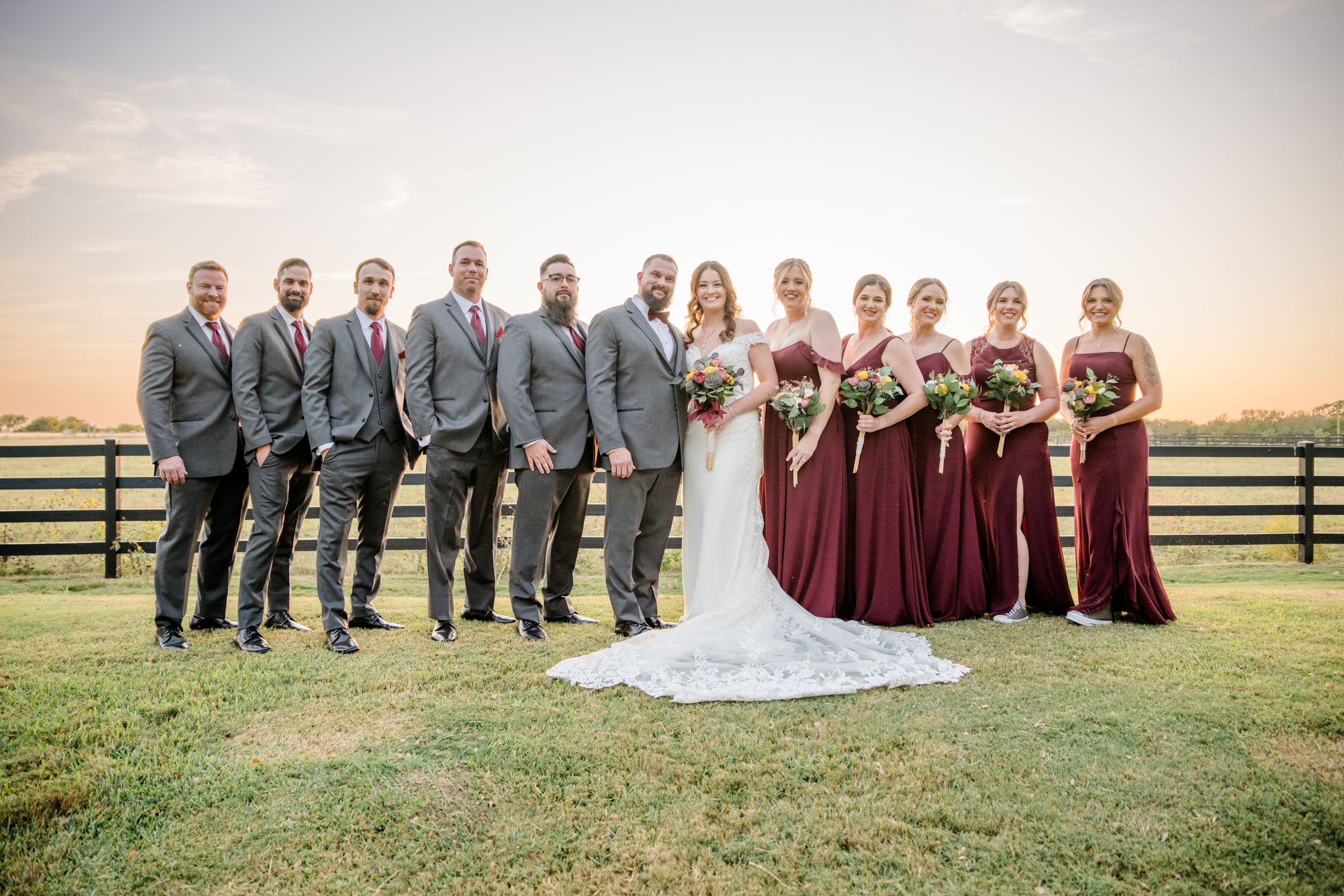 Wedding party poses in burgundy dresses and gray suits at sunset on a grass field with wooden fence backdrop.