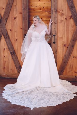 A bride poses in an elegant white lace wedding gown against a rustic wooden barn backdrop.