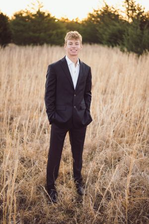 A young man in a dark suit stands in a field of tall grass at sunset.