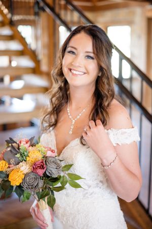 Beautiful bride in off-shoulder lace gown holds colorful autumn bouquet with yellow and pink roses in rustic indoor setting.