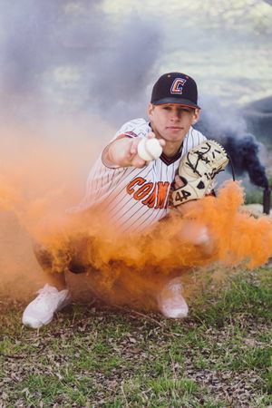 Baseball player in white uniform crouches with orange smoke effect in dramatic sports portrait.