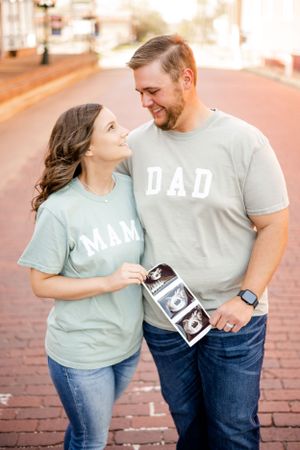 Expectant couple wearing 'Mom' and 'Dad' shirts hold ultrasound photo on brick sidewalk.