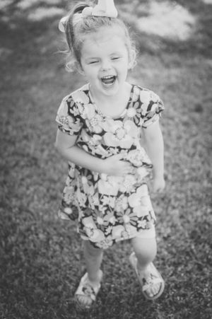 Black and white photo of young child laughing in floral dress with bow in hair.