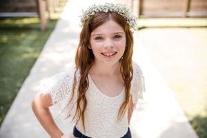Young girl in white lace dress and flower crown poses outdoors in sunlight.