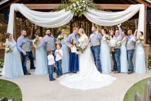 Wedding party poses under elegant white draped arch with floral decor during outdoor ceremony.