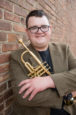 Musician in glasses and blazer poses with golden trumpet against brick wall backdrop.