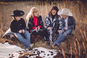 Group wearing western attire sits on cowhide blanket in rustic field setting.